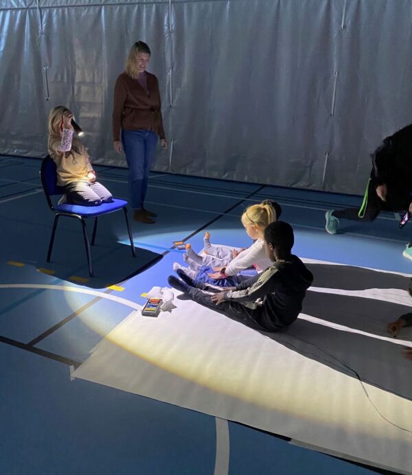 Et barn sitter på en stol i gymsalen i et mørkt rom og leker med skyggen til to barn som sitter på gulvet i gymsalen. Foto