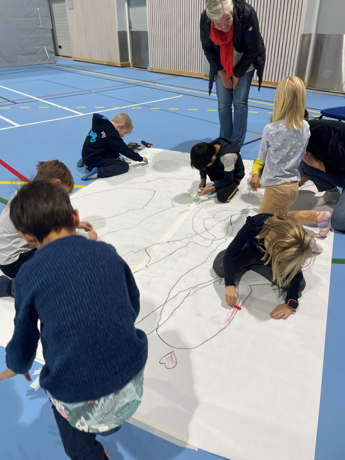 Barn sitter på gulvet i en gymsal og tegner på et stort lerret, en voksen står over og ser på tegningene. Foto.