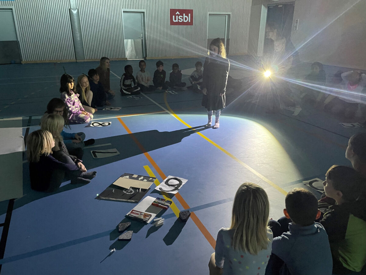 Barn sitter på gulvet i en gymsal i ring, med et barn som leker med skyggen i midten av ringen. Foto