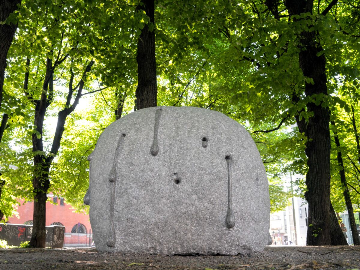 Steinskulptur står utendørs under grønne trær. Foto.