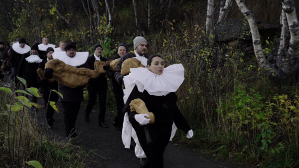 En prosesjon av personer på linje i skogen kledt i gammeldagse prestekjortler. Stillbilde fra film