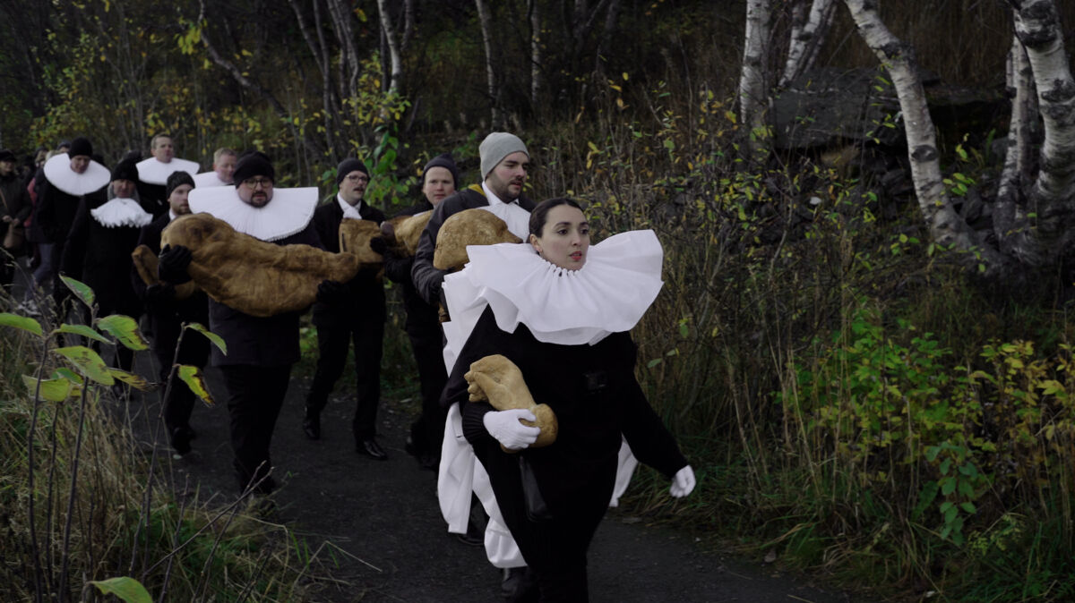 En prosesjon av personer på linje i skogen kledt i gammeldagse prestekjortler. Stillbilde fra film