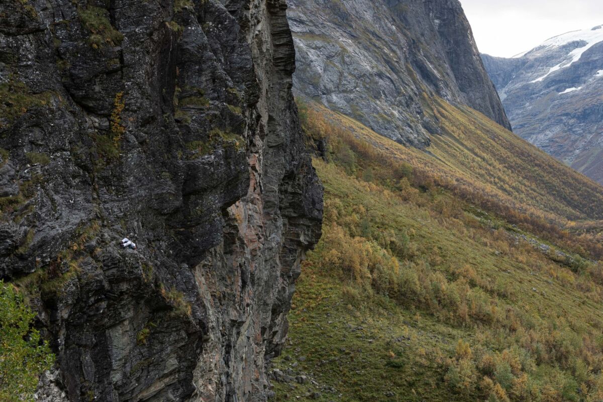 Klatrer ligger høyt oppe i en fjellvegg, med fjellandskap i bakgrunnen. Stillbilde fra film.