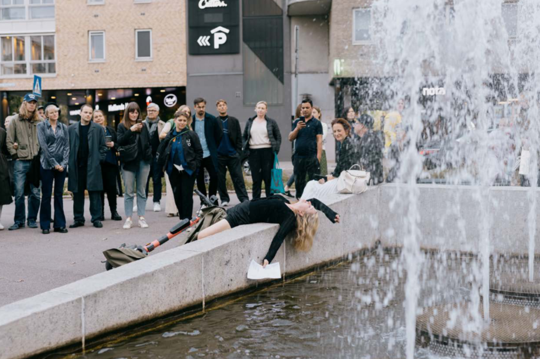 Kunstperformance med kunstner halvt liggende i fontene, med publikum i bakgrunnen. Foto