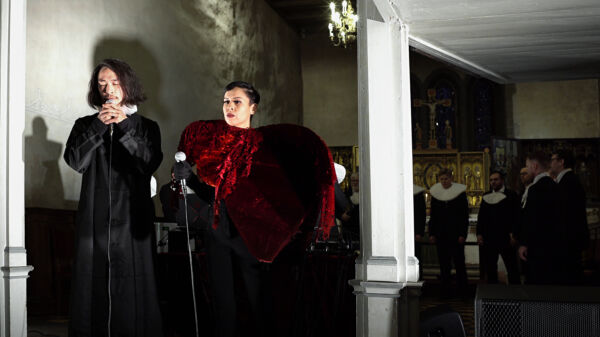 To personer i et kirkerom. Den ene holder og synger i en mikrofon. Foto.