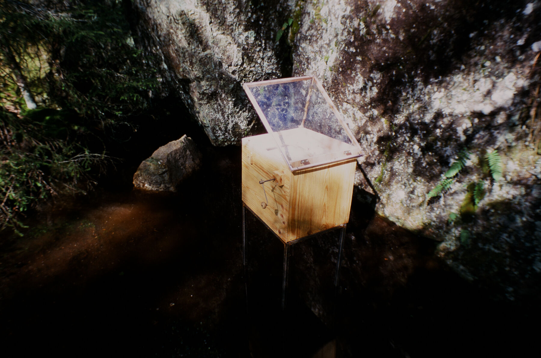 En trekasse som ligner en lirekasse står i en bekk mot en steinvegg. Foto.