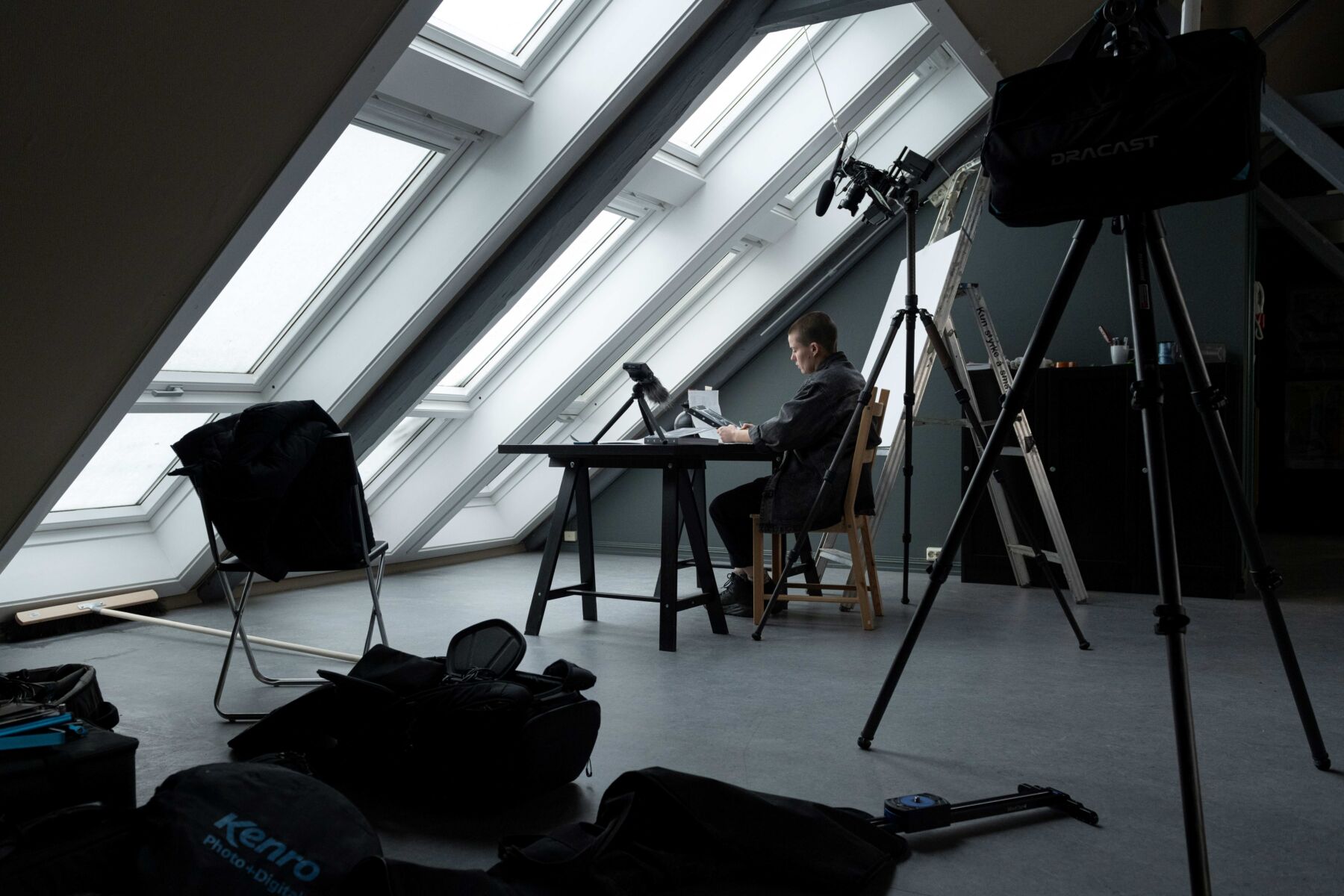 En person sitter i et rom med skråtak og -vinduer. Rundt står utstyr til filmproduksjon. Foto.