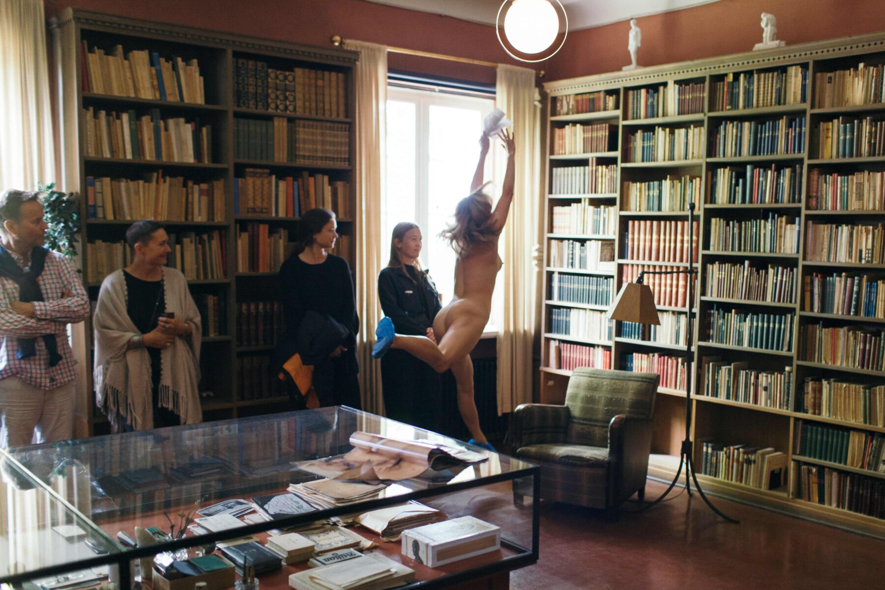 Naken person danser i et bibliotek. Fire mennesker står inntil veggen og betrakter den dansende. Foto.