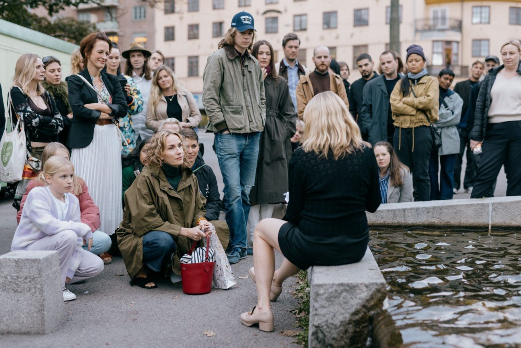 Kunstperformance sitter på kanten av et lite fontenebasseng. Publikum i bakgrunnen. Foto.