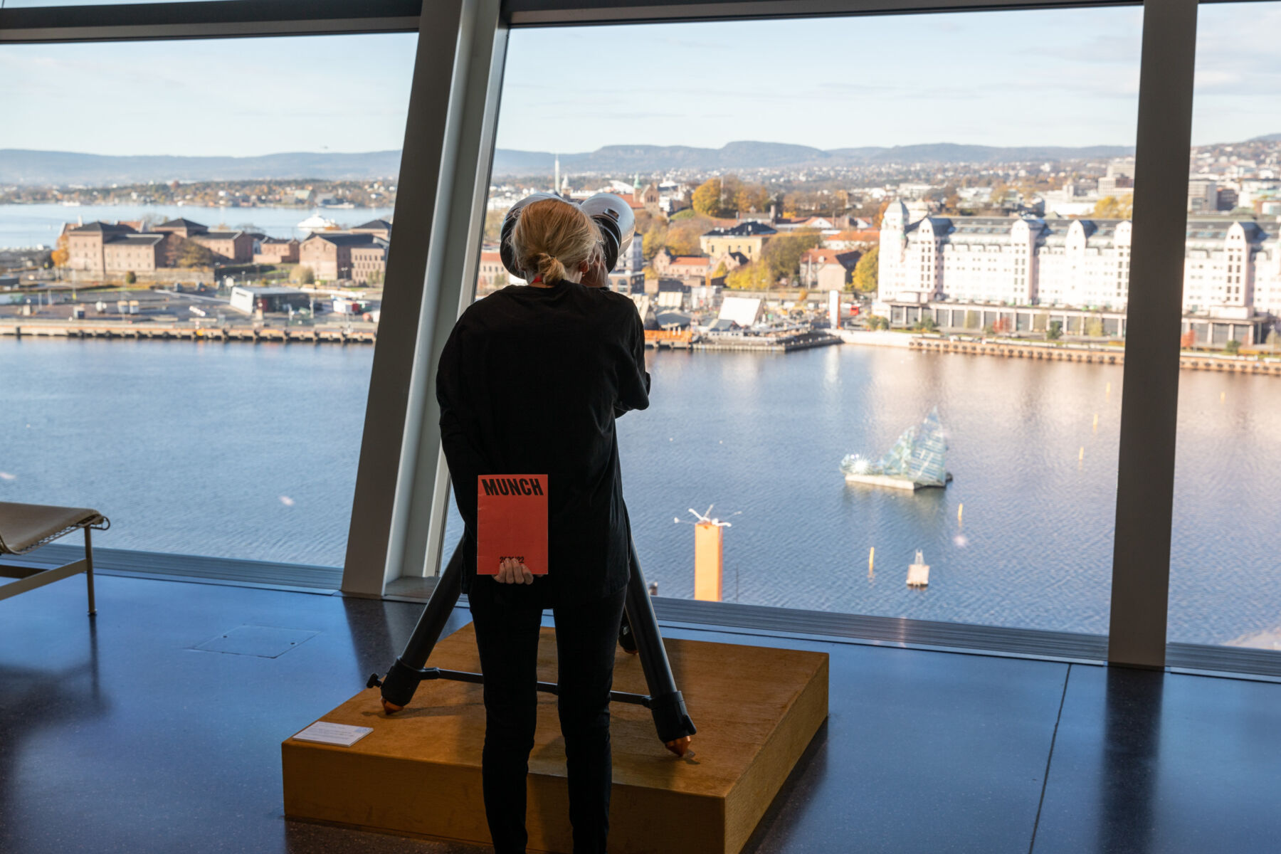 Person med ryggen til står og ser gjennom en kikkert på utsikten over fjorden og byen. Foto.