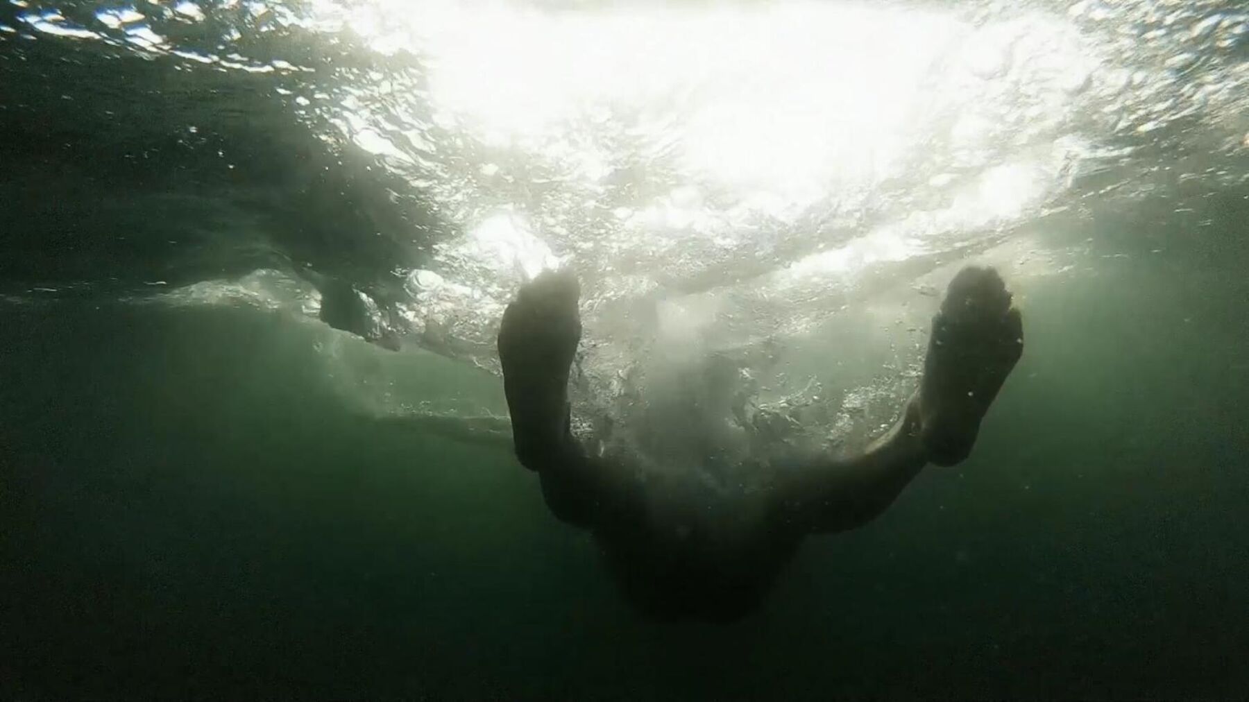 Person liggende i vann, tatt under ifra, med sol som skinner gjennom vannet. Stillbilde.