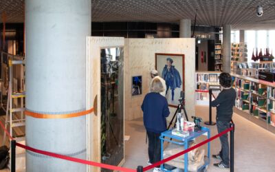 To personer står i et bibliotek og ser på en tredje person som studerer kunst hengt opp på vegger. Foto.