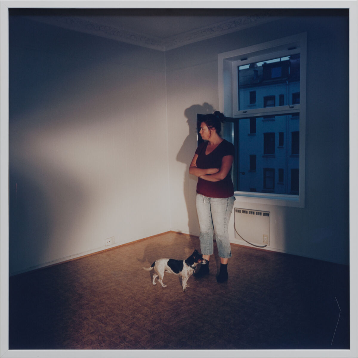 Person og hund står på et gulv i en leilighet. I bakgrunnen er et vindu med utsikt til en bygård. Foto.