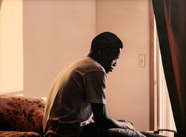 Mann sitter på en seng og ser ned på gulvet. Foto.