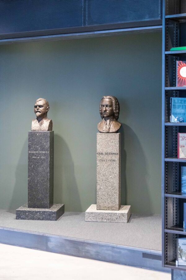 To byster på hver sin sokkel står ved siden av hverandre foran en mørkegrønn vegg. Foto.