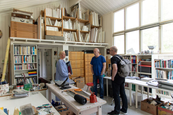 Tre personer står i et stort studio omringet av materialer og bøker.