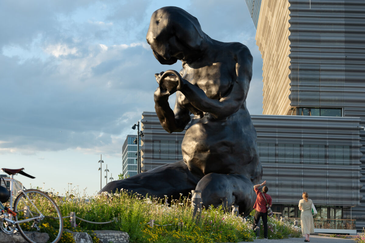 A monumental bronze placed on a meadow in front of MUNCH museum. Two peolpe looking up at the sculpture. Photo