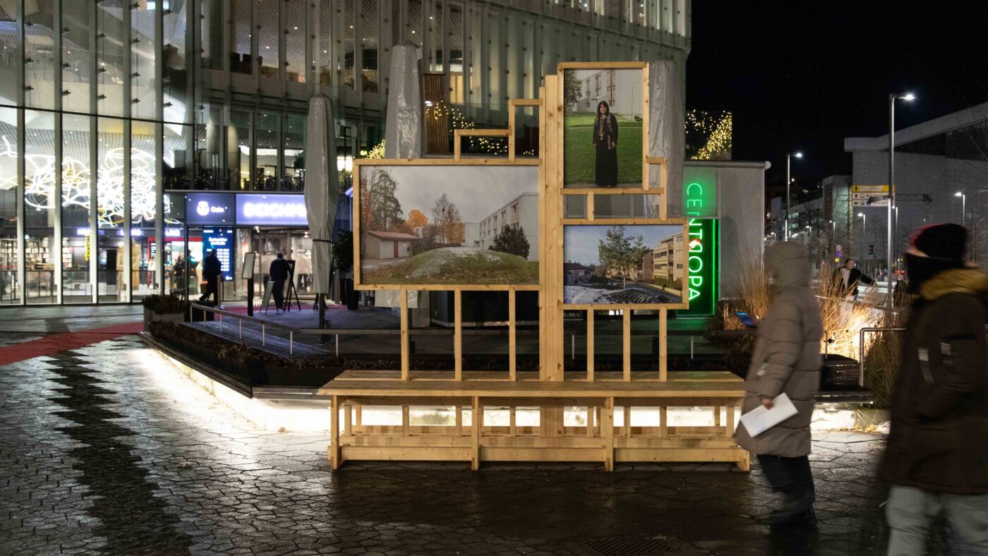 Kveldsbilde av utemiljø foran Deichman Bjørvika, fotografier er utstilt på en trestruktur foran biblioteksbygningen, to anonyme mennesker passerer i forgrunn. Foto