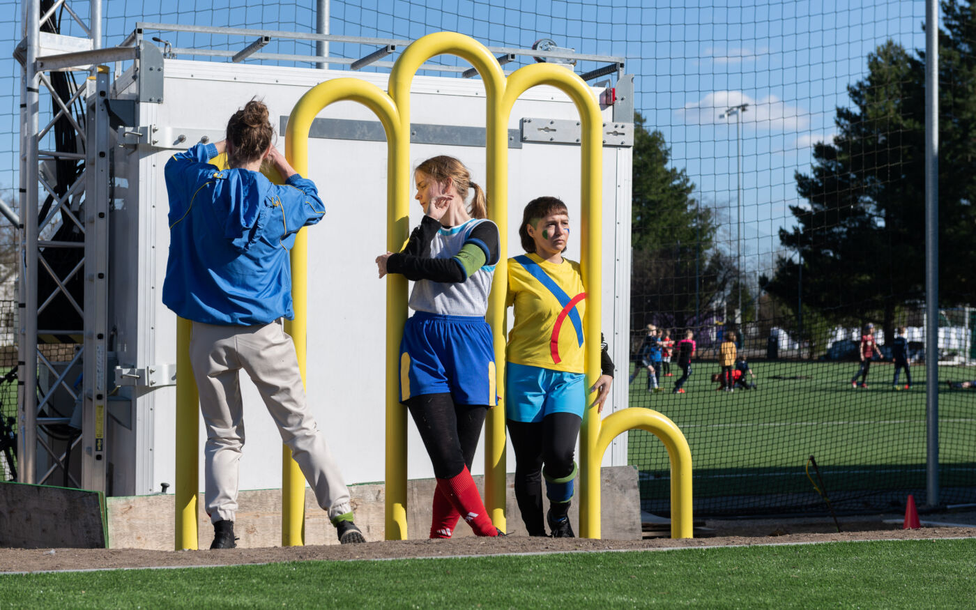 Tre mennesker utfører en performance ved en gul bueformet skulptur, plassert ved en fotballbane. Foto