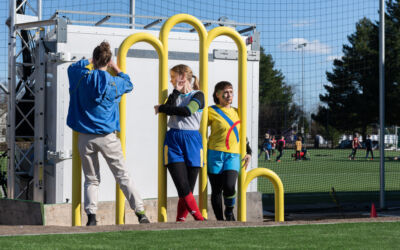 Tre mennesker utfører en performance ved en gul bueformet skulptur, plassert ved en fotballbane. Foto
