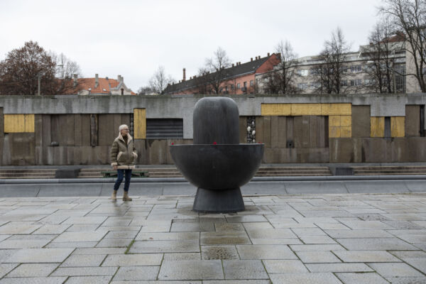 Steinlagt plass, med steinskulptur og person midt i bildet. Steinmur med innslag av gull i bakgrunnen. Foto