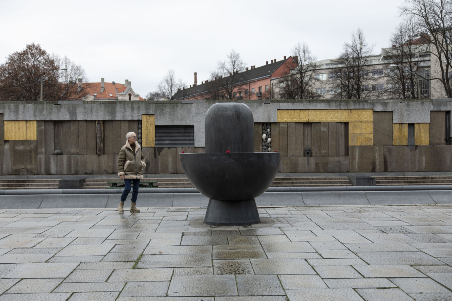 Steinlagt plass, med steinskulptur og person midt i bildet. Steinmur med innslag av gull i bakgrunnen. Foto