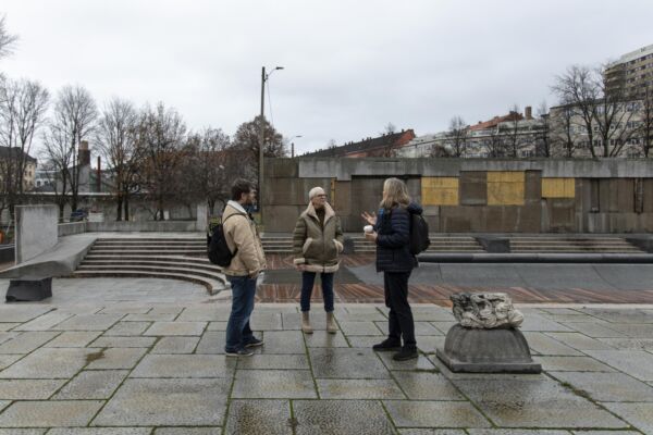 Tre personer står å prater på en steinbelagt plass, murvegg med innslag av gull i bakre del av bildet. Foto