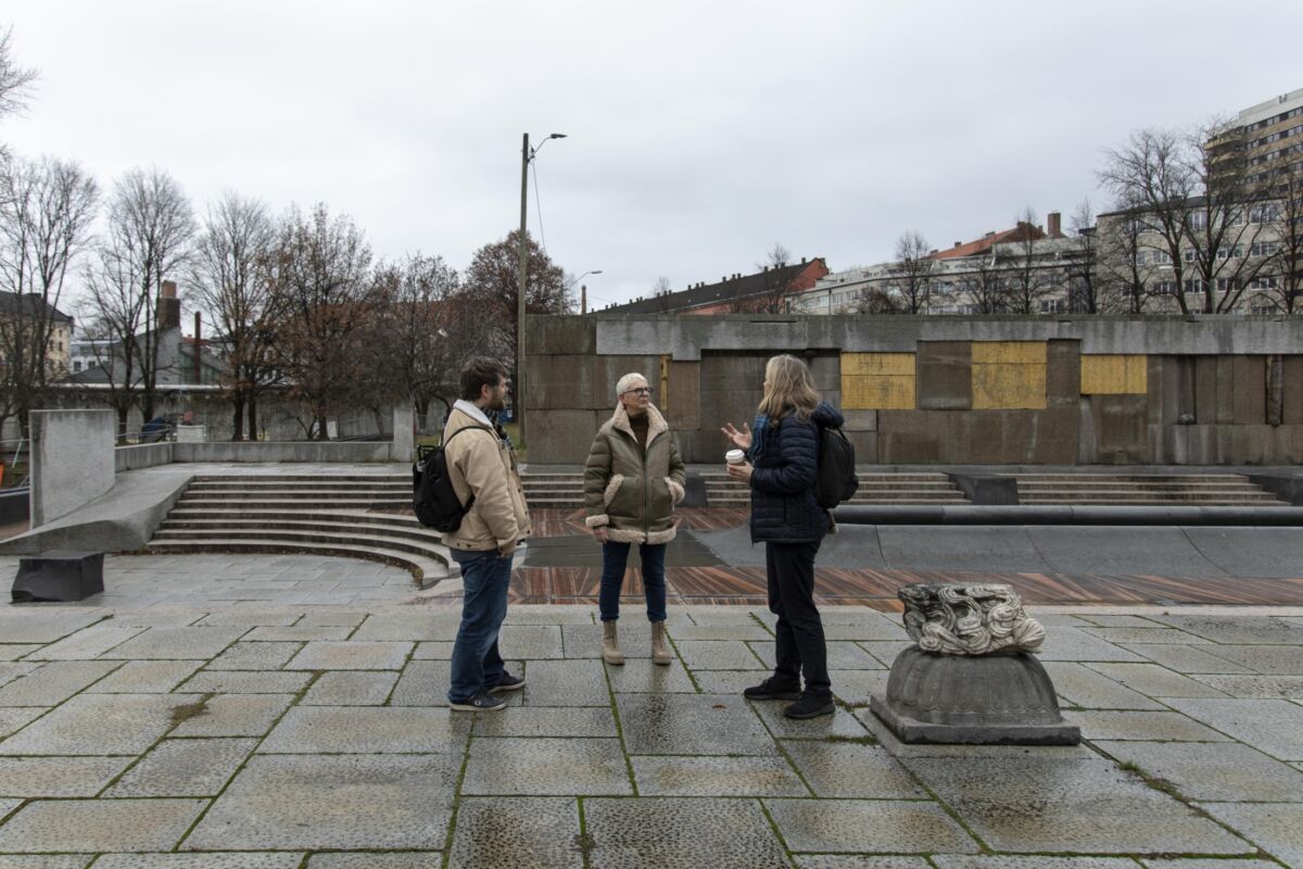 Tre personer står å prater på en steinbelagt plass, murvegg med innslag av gull i bakre del av bildet. Foto