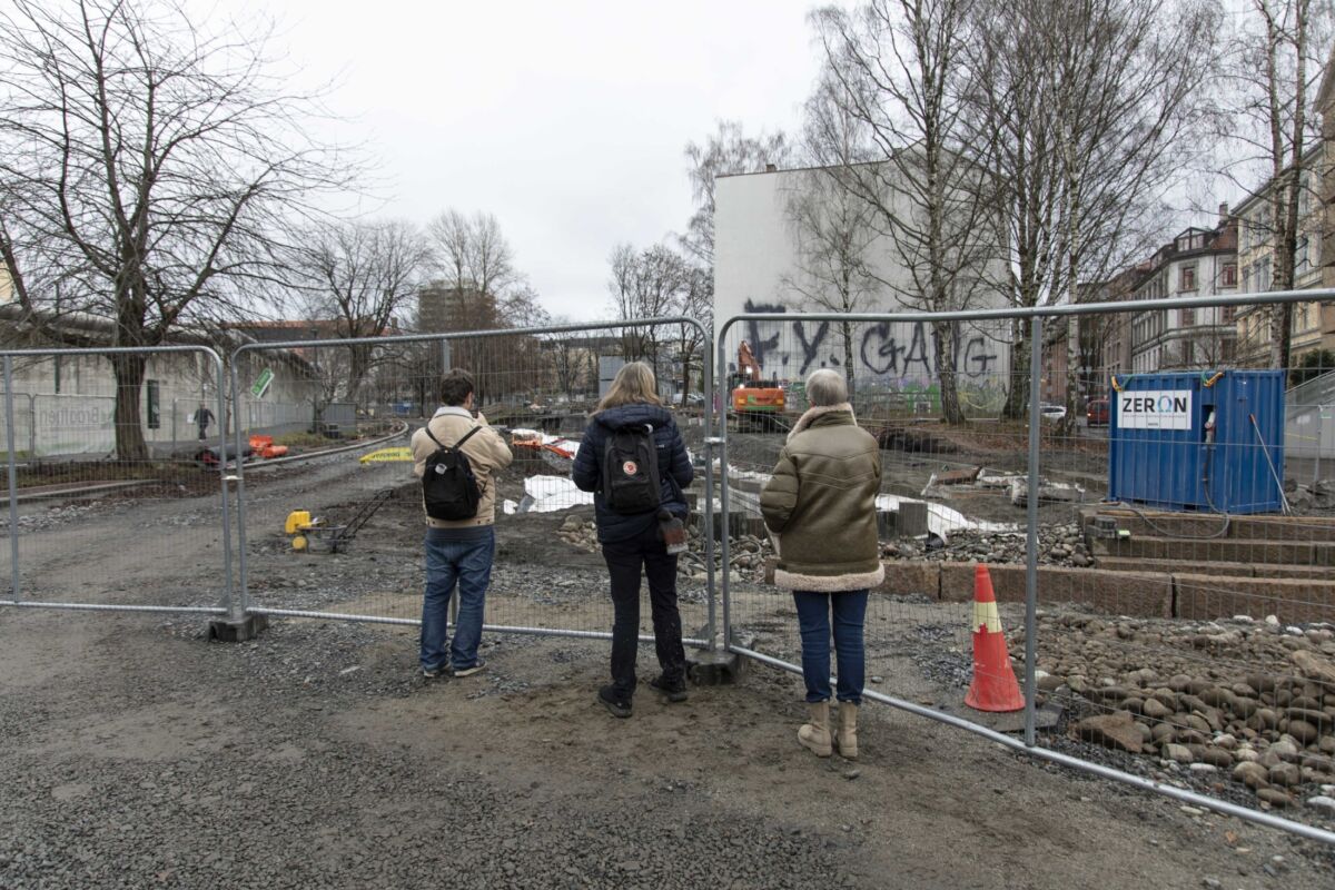 Tre personer står å ser gjennom et byggegjerde på en anleggsplass. Foto