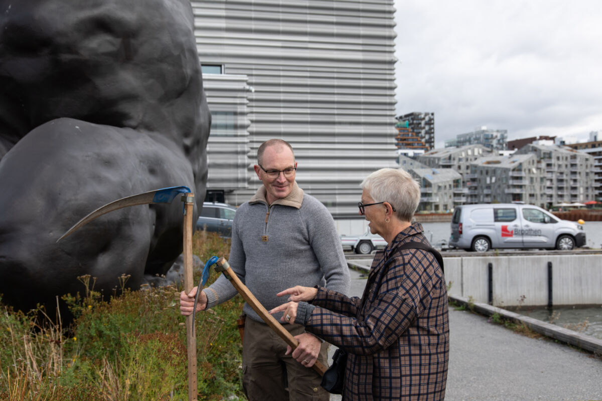 To personer med ljå ved et lite eng parti, utsnitt av bronseskulptur og bygg i bakgrunnen. Foto