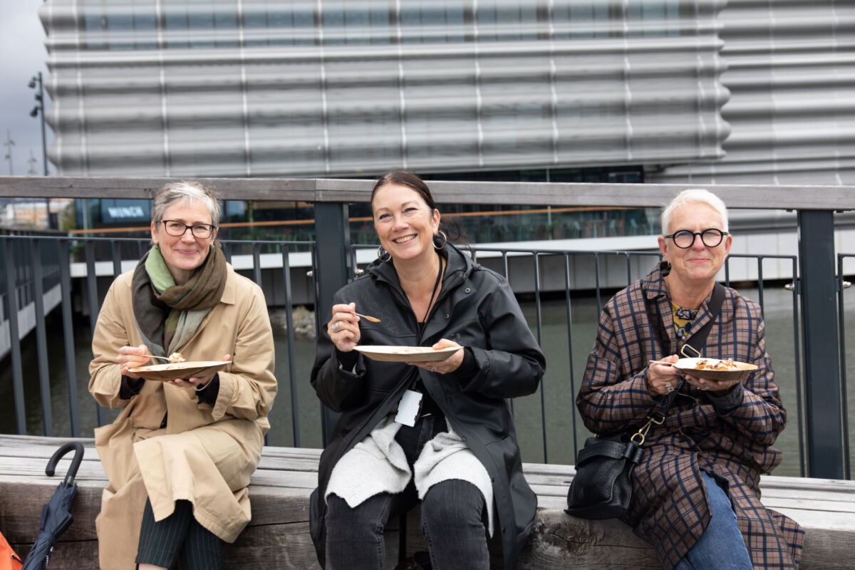 Tre personer sitter på en benk foran MUNCH museet. De spiser og smiler til kamera. Foto