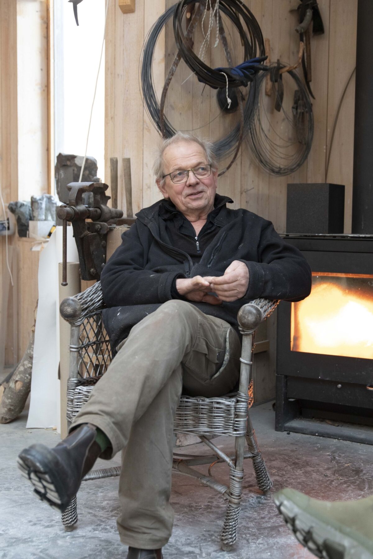 Kunstner Kristian Blystad sitter i stol foran peis i sitt atelier. Foto