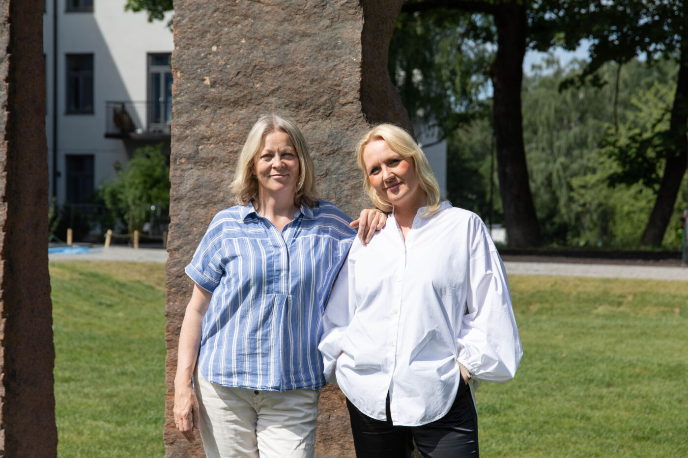 To mennesker ser i kamera, steinskulptur og gressslette i bakgrunnen. Foto
