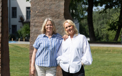 To mennesker ser i kamera, steinskulptur og gressslette i bakgrunnen. Foto