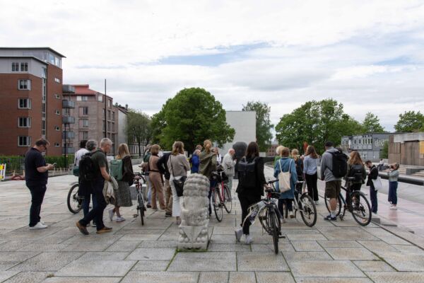Mange mennesker står samlet rundt en guide på en plass. Foto.