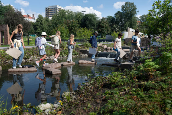 Foto av flere barn og ungdom som krysser steiner over en bekk i Klosterenga park.
