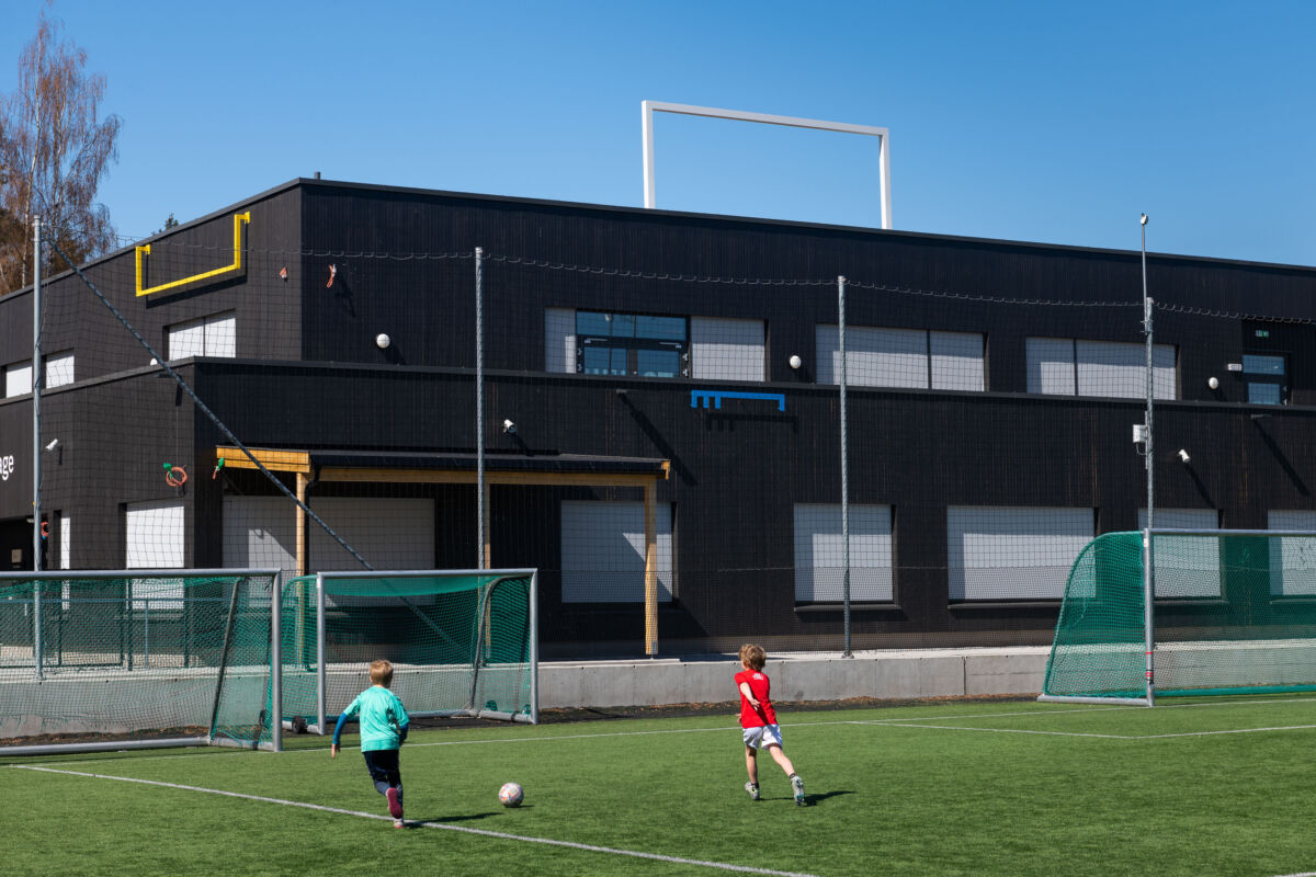 To barn spiller fotball på en gressplen foran et stort, mørkebrunt bygg. Foto.