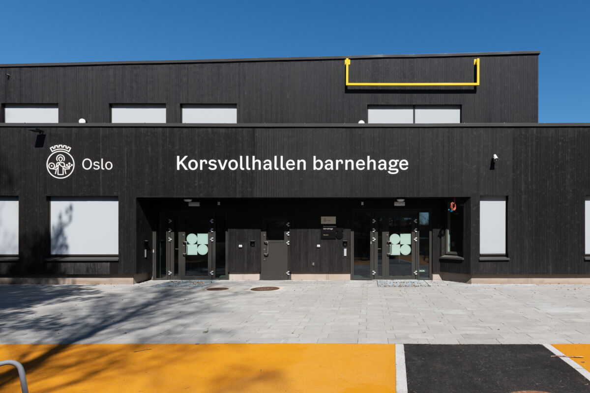 Bygning med teksten "Korsvoll barnehage" påskrevet. En gul skulptur med rette vinkler henger fra veggen. Foto.