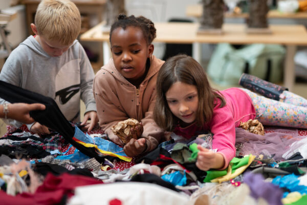 Tre barn lener seg over et bord med tekstil. Foto.