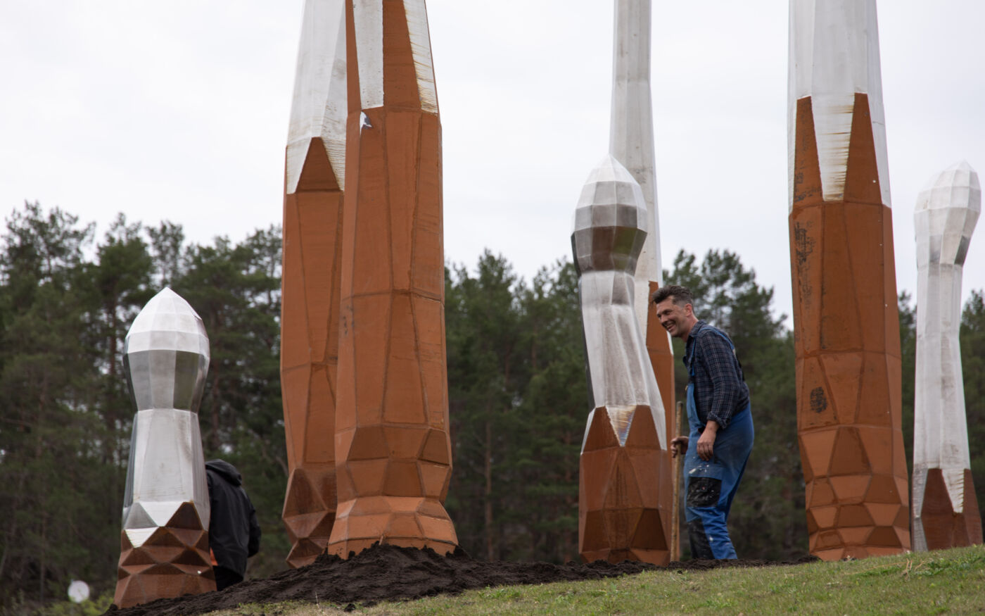 Person står blant en skulpturgruppe. Skulpturene er spirelignende former som strekker seg fra bakken og opp mot himmelen. Foto.