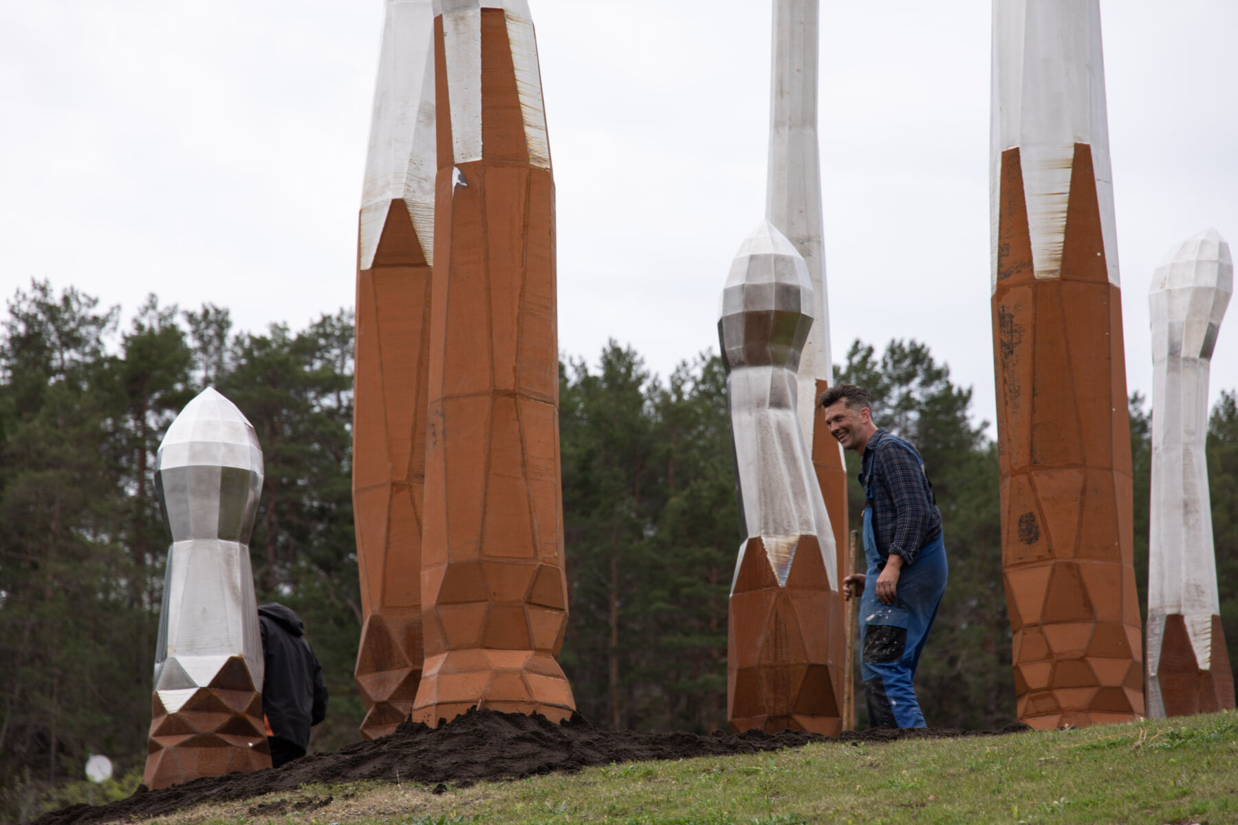 Person står blant en skulpturgruppe. Skulpturene er spirelignende former som strekker seg fra bakken og opp mot himmelen. Foto.