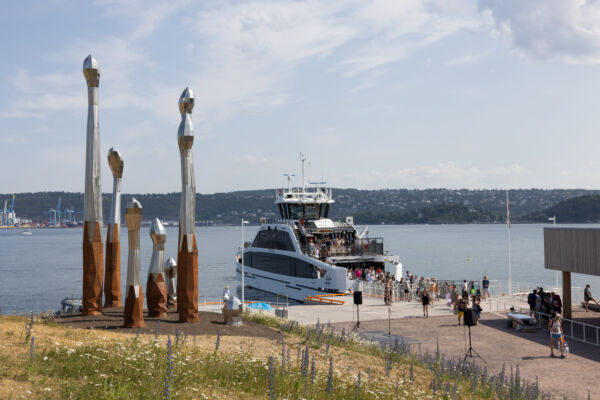Kunstverk stående på en brygge. En båt i bakgrunnen. Foto.