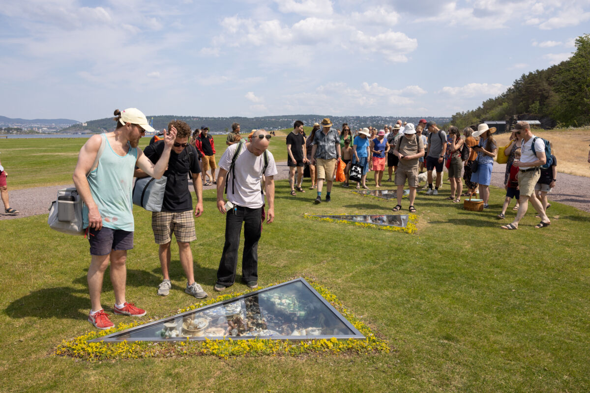 Mennesker ser på nedsenket skulptur i grønt landskap. Foto.