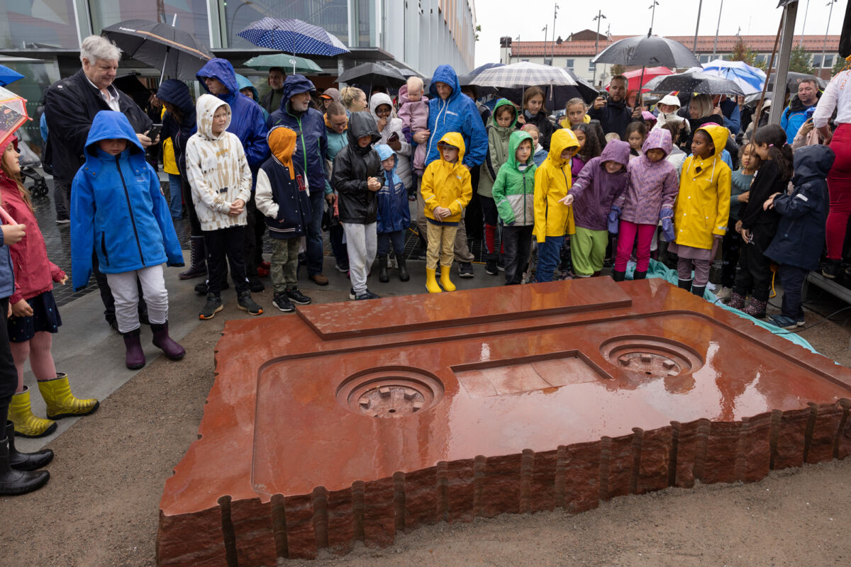En stor gruppe mennesker står rundt kunstverk; steinskulptur utformet som kassett. Foto