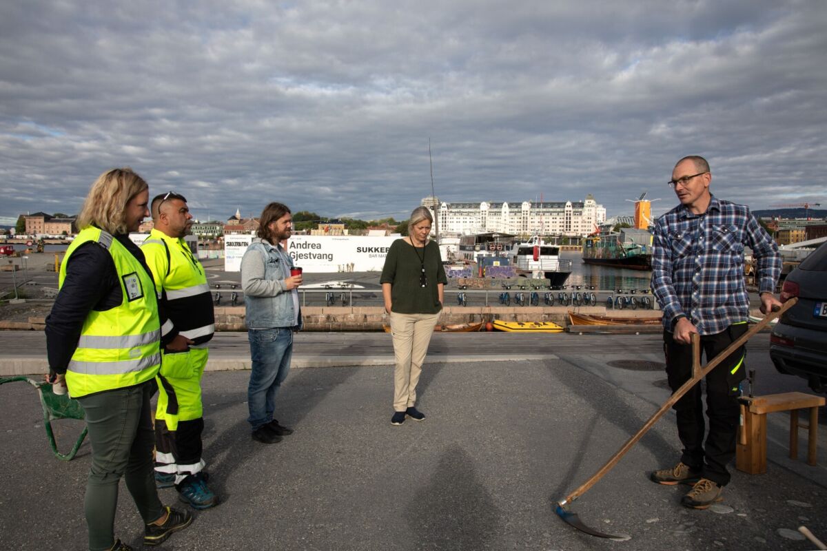 En mann holder en ljå, fire personer står rundt og ser på. Foto.