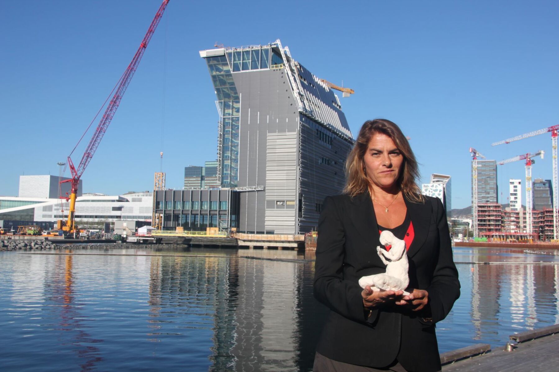 Tracey Emin ser i kamera, hun holder en liten skulptur i armene, fjord og MUNCH museet i bakgrunnen. Foto