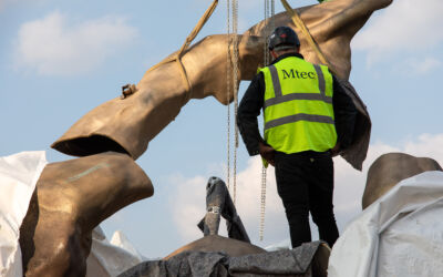 En mann i gul vest står å ser på  at deler av en stor bronseskulptur løftes. Foto