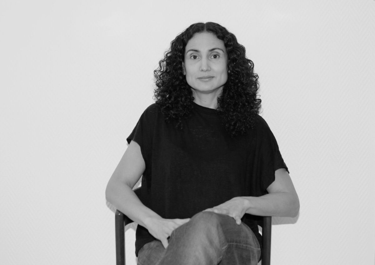 Portrettbilde i svart-hvitt av en person som sitter på en stol. Personen har langt, bølgete mørkt hår og mørk t-skjorte. Personen ser rett i kamera. Foto.