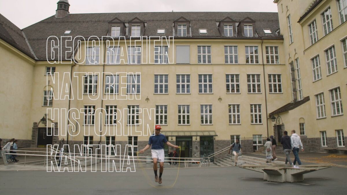 En person hopper hoppetau i en skolegård. Skolebygningen i bakgrunnen er gul og stor med mange vinduer. Stillbilde fra film.
