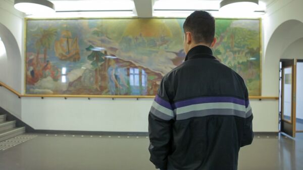 Person står med ryggen til kamera, i bakgrunnen er et bredt, innrammet veggmaleri med lyse farger. Stillbilde fra film.